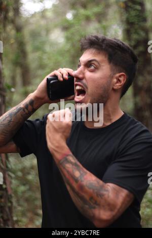 Un giovane frustrato urla nel suo cellulare durante un'intensa telefonata, in piedi in mezzo a una foresta pacifica Foto Stock