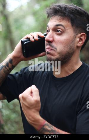 Un giovane frustrato urla nel suo cellulare durante un'intensa telefonata, in piedi in mezzo a una foresta pacifica Foto Stock