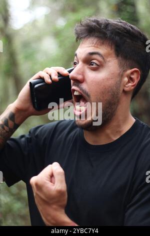 Un giovane frustrato urla nel suo cellulare durante un'intensa telefonata, in piedi in mezzo a una foresta pacifica Foto Stock