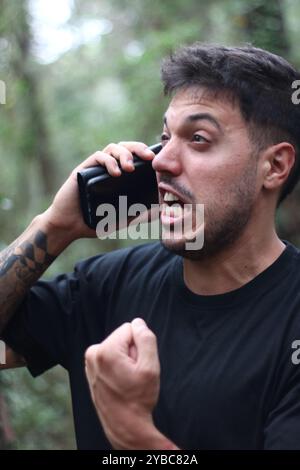 Un giovane frustrato urla nel suo cellulare durante un'intensa telefonata, in piedi in mezzo a una foresta pacifica Foto Stock