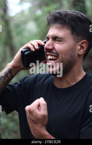 Un giovane frustrato urla nel suo cellulare durante un'intensa telefonata, in piedi in mezzo a una foresta pacifica Foto Stock