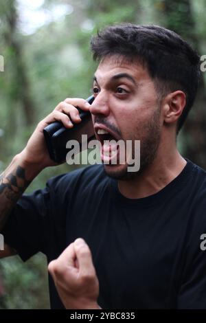 Un giovane frustrato urla nel suo cellulare durante un'intensa telefonata, in piedi in mezzo a una foresta pacifica Foto Stock