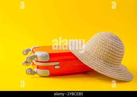Valigia da viaggio in miniatura rossa sdraiata con un cappello di paglia sulla parte superiore, su uno sfondo giallo e concetto di viaggio Foto Stock