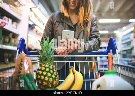 Donna, telefono e corridoio del supermercato per acquisti, coupon digitali e offerte di merchandising. Persona femminile, vendita e negozio per opzioni in offerta, vendita al dettaglio Foto Stock