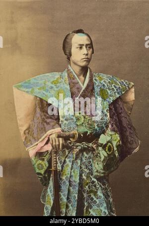 Guerriero giapponese, 1868-1880. Ritratto in studio di un uomo in abiti da samurai tradizionali. Indossa un kamishimo elaborato e porta due spade. Una delle spade è infilata nella sua cintura e l'altra tiene in posizione eretta con la mano appoggiata sul pomelo. Foto Stock