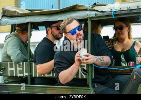 Un vivace gruppo di amici si riunisce su una jeep in stile militare, ridendo e condividendo bevande in una giornata di sole, creando un'atmosfera rilassata e festosa Foto Stock