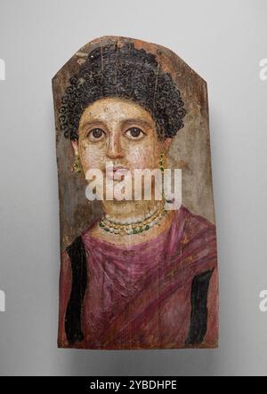 Mummy Portrait, d.C. 75-100. Ulteriori informazioni: Questo ritratto di una donna presenta molte delle caratteristiche distintive dei pannelli funerari romano-egiziani. Lo stile dei suoi capelli scuri e ricci, legati in un panino sciolto in cima alla testa, era di moda durante il periodo Flavio (68-96) e aiuta a datare il ritratto agli anni intorno a quel periodo. Il viso della donna è dipinto in tonalità carne e salmone, lo sfondo è grigio scuro e il suo indumento è rosato-lavanda (tradizionale per i ritratti funerari femminili) con clavi neri (strisce intrecciate) che corrono verticalmente lungo la parte anteriore. Lei Foto Stock