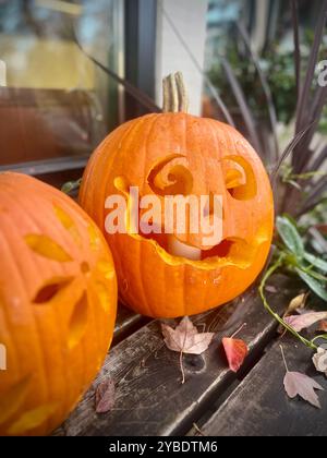 Una zucca intagliata Jack-o'-lanterna con un design giocoso si trova su una mostra autunnale, circondata da foglie autunnali, che celebrano la stagione di Halloween Foto Stock