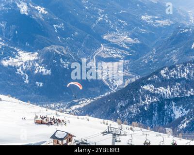 Un parapendio galleggia sopra le piste innevate di Brianza, in Francia, con una valle panoramica e strade tortuose visibili sotto. La gente si riunisce vicino a uno sci Foto Stock