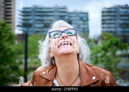 Felice donna caucasica anziana con capelli grigi in piedi che guarda ridere di fronte al giardino della città. Foto Stock