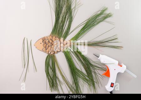 attività didattica dell'ora prescolare, giocattolo per pesci fatto di aghi a cono di pino, processo creativo, pistola per colla a caldo, sfondo grigio, vista dall'alto verso il basso. immagine di Foto Stock