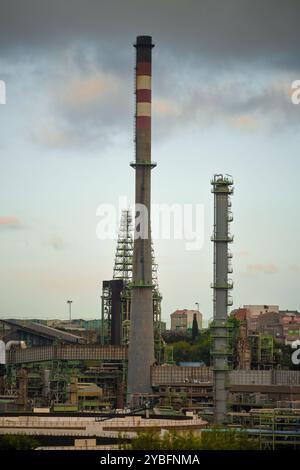 Un teleobiettivo di un paesaggio industriale con due alti fumogeni e complesse strutture di raffineria, circondate da edifici urbani. Cielo coperto aggiungi Foto Stock