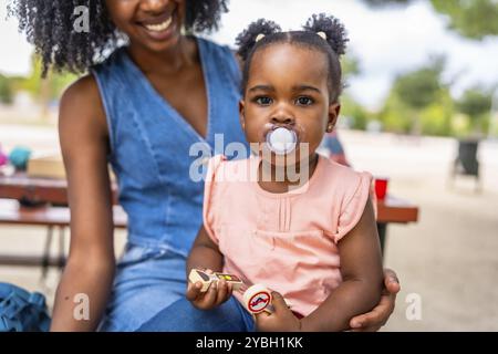 Graziosa ragazza africana con succhietto seduto in grembo alla mamma nel parco Foto Stock