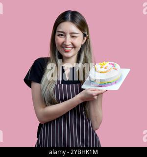 Giovane donna asiatica con un grembiule in stato festivo o festivo che tiene una torta colorata con vari spruzzi sul condimento. Ritratto su backgroun rosa Foto Stock