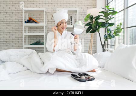 Giovane donna asiatica con un sorriso, siediti su un letto applicando il mascara, tenendo uno specchio, indossando un accappatoio e un asciugamano sulla testa. Foto Stock