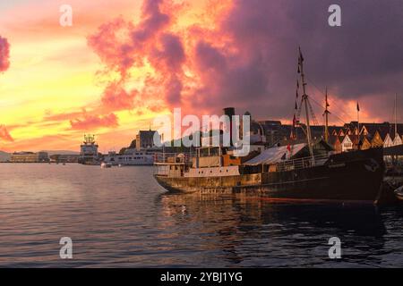 7 settembre 2024 Bergen, Norvegia Sun tramonta su una vecchia barca a vapore ormeggiata di fronte ai colorati magazzini convertiti sul porto di Foto Stock