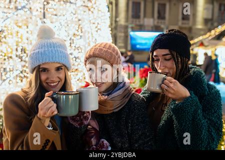 Gruppo multiculturale di amiche che bevono bevande calde in un mercatino di Natale. Un'amicizia variegata, moda invernale e festeggiamenti natalizi Foto Stock