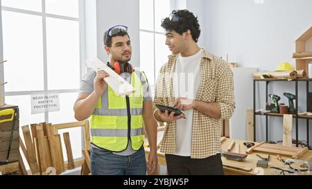 Due uomini si consultano su un progetto in un'officina di falegnameria luminosa, circondata da utensili per la lavorazione del legno e attrezzature di sicurezza. Foto Stock