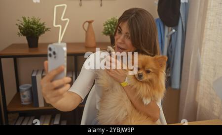 Una giovane donna ispanica a casa si fa un selfie con il suo cane pomerania al chiuso, incarnando un intimo momento di vita. Foto Stock