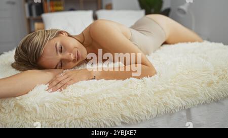 Bella donna bionda che si rilassa su un comodo letto in una camera da letto interna di casa, sdraiata a occhi chiusi e con un'espressione pacifica. Foto Stock