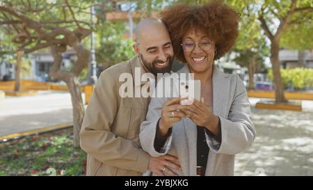Una coppia eterogenea sorride con gioia mentre si fanno un selfie insieme in un parco urbano, mettendo in risalto affetto, tecnologia e tempo libero. Foto Stock