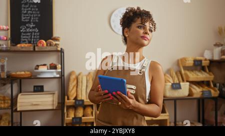Giovane donna ispanica in piedi in una panetteria con un tablet, circondata da pani di pane e dolci, che ha un aspetto premuroso. Foto Stock