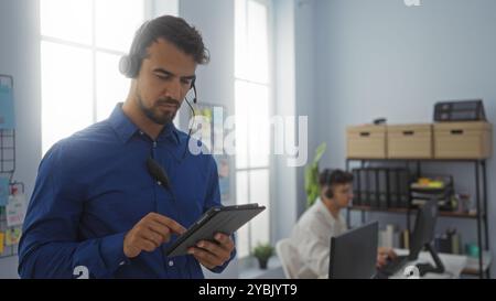 Uomini ispanici che lavorano come dipendenti utilizzando le cuffie in un ufficio, con un uomo che si concentra su un tablet e l'altro su un computer. Foto Stock