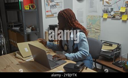 Una detective afroamericana concentrata lavora sui fascicoli di un caso in un ufficio disordinato della stazione di polizia Foto Stock