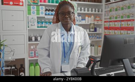 Una giovane farmacista afroamericana si trova con fiducia in una moderna farmacia interna dietro il bancone. Foto Stock