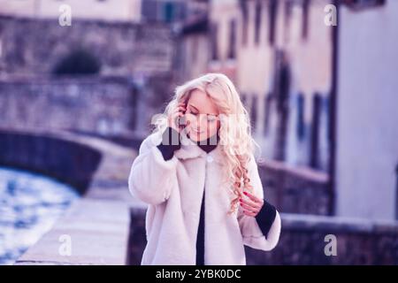 Una giovane donna passeggia felicemente lungo un canale in europa, chiacchierando sul suo smartphone in una giornata invernale di sole. Trasuda eleganza in un cappotto di pelliccia, godendosi la fr Foto Stock