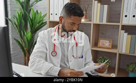 Un giovane uomo di colore adulto con una barba che indossa un camice da laboratorio esamina i documenti in un ufficio clinico pieno di luce. Foto Stock