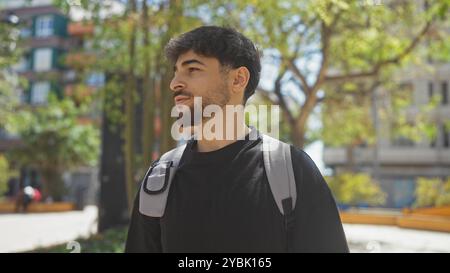 Un giovane con la barba cammina con cura attraverso un parco urbano illuminato dal sole, incarnando un comportamento informale ed elegante. Foto Stock
