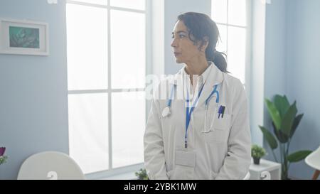 Una donna sicura di sé medico si trova contemplativamente in una luminosa stanza d'ospedale, trasudando professionalità ed esperienza. Foto Stock