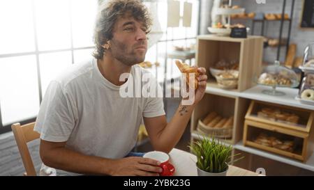 Giovane che si gusta un croissant in un ambiente da forno interno con vari dolci, seduto a un tavolo mentre tiene una tazza di caffè. Foto Stock