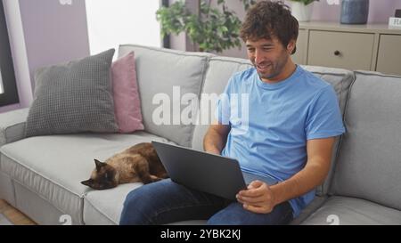 Un giovane sorridente in un abbigliamento casual lavora sul suo computer portatile mentre si siede su un divano grigio accanto al suo gatto siamese riposante in un accogliente soggiorno. Foto Stock