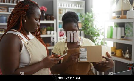 Due donne afroamericane che lavorano in un negozio di arredamento per la casa, una con una scatola e l'altra con uno smartphone, circondate da vari oggetti d'arredo Foto Stock