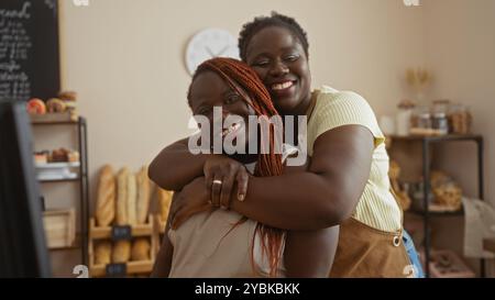 Due donne afroamericane che sorridono e si abbracciano in un accogliente negozio interno di panetteria con pane fresco e pasticcini sullo sfondo Foto Stock