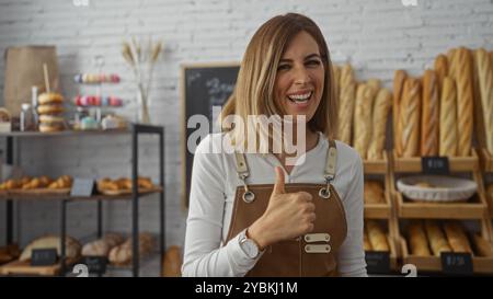 Donna bionda sorridente e con il pollice in una panetteria, circondata da una varietà di pane fresco, pasticcini e bagel, creando un accogliente indoo Foto Stock