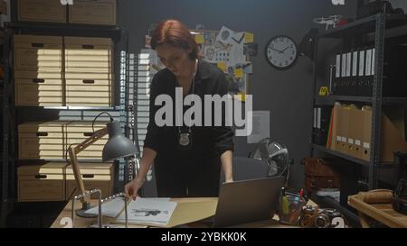 Detective con la testa rossa che lavora in una stazione di polizia disordinata, analizzando i documenti a tarda notte. Foto Stock