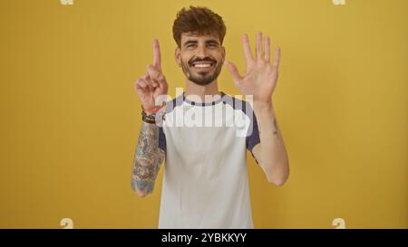 Bellissimo giovane con barba e tatuaggi che fa un segno numero sei con le dita su uno sfondo giallo. Foto Stock