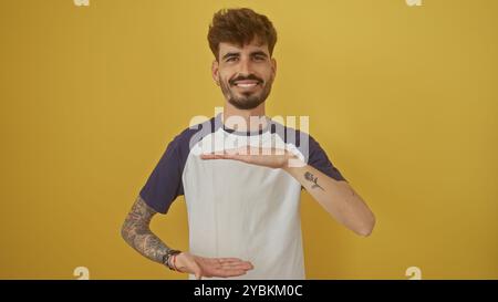 Un giovane ispanico sorridente con barba e tatuaggi, con indosso una t-shirt casual, fa gesti con le mani su uno sfondo giallo vibrante. Foto Stock