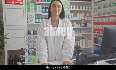 Una giovane farmacista ispanica sorridente si trova con fiducia in un cappotto bianco all'interno di una farmacia ben fornita Foto Stock