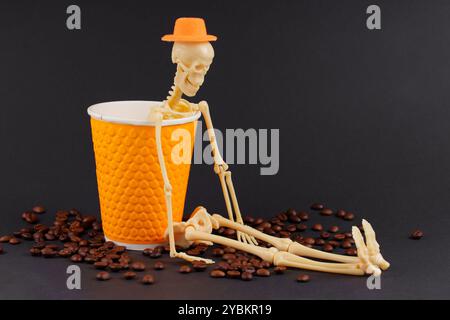 Scheletro accanto alla tazza di caffè arancione brillante con chicchi di caffè sparsi su una superficie scura. Decorazione della caffetteria Halloween. Foto Stock