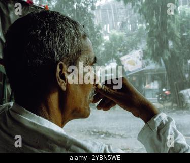 Un vecchio seduto sulla sedia e perso nei pensieri mentre guardavano la pioggia fuori in Bangladesh. Una fotografia di strada di un uomo solo. Foto Stock