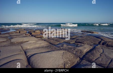 Il promontorio roccioso di Point Cartwright si protende verso il mare con onde da surf che si infrangono su di esso. La superficie liscia delle rocce rende facile camminare fino all'ex Foto Stock