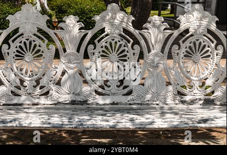 Dettagli della panca da giardino in ghisa dipinta di bianco in stile vittoriano nel parco cittadino. Luce solare sulla superficie dello schienale di un banco in ghisa antica decorata vuota in Foto Stock