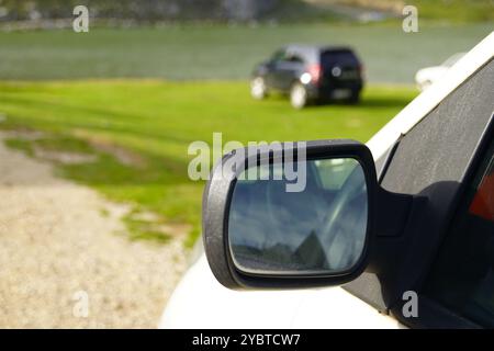 Una foto ravvicinata di uno specchio retrovisore contro una vista sfocata di un lago di montagna e di un SUV nero parcheggiato su un prato verde. Viaggio in Montenegro Foto Stock