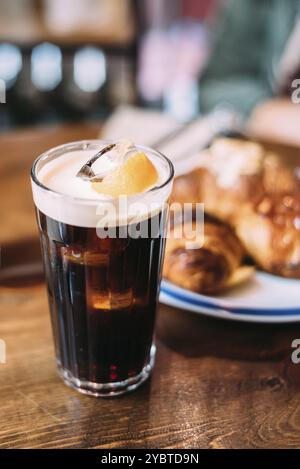 Caffè ghiacciato in un alto bicchiere con panna versata su un vecchio tavolo rustico in legno con pasticcini sfocati sullo sfondo. Drink estivo freddo con spazio fotocopie Foto Stock