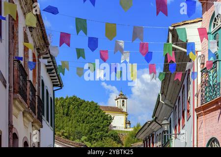 Strada decorata con bandiere per i festeggiamenti di San Giovanni con case in stile coloniale e una chiesa sullo sfondo nella città di Ouro Preto Foto Stock