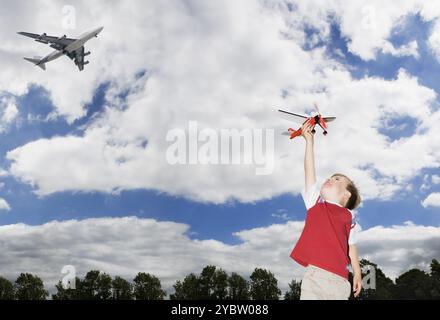 Giovane ragazzo caucasico che gioca con un aeroplano giocattolo mentre un aereo passeggeri vola sopra temi di ispirazione futura per l'immaginazione Foto Stock